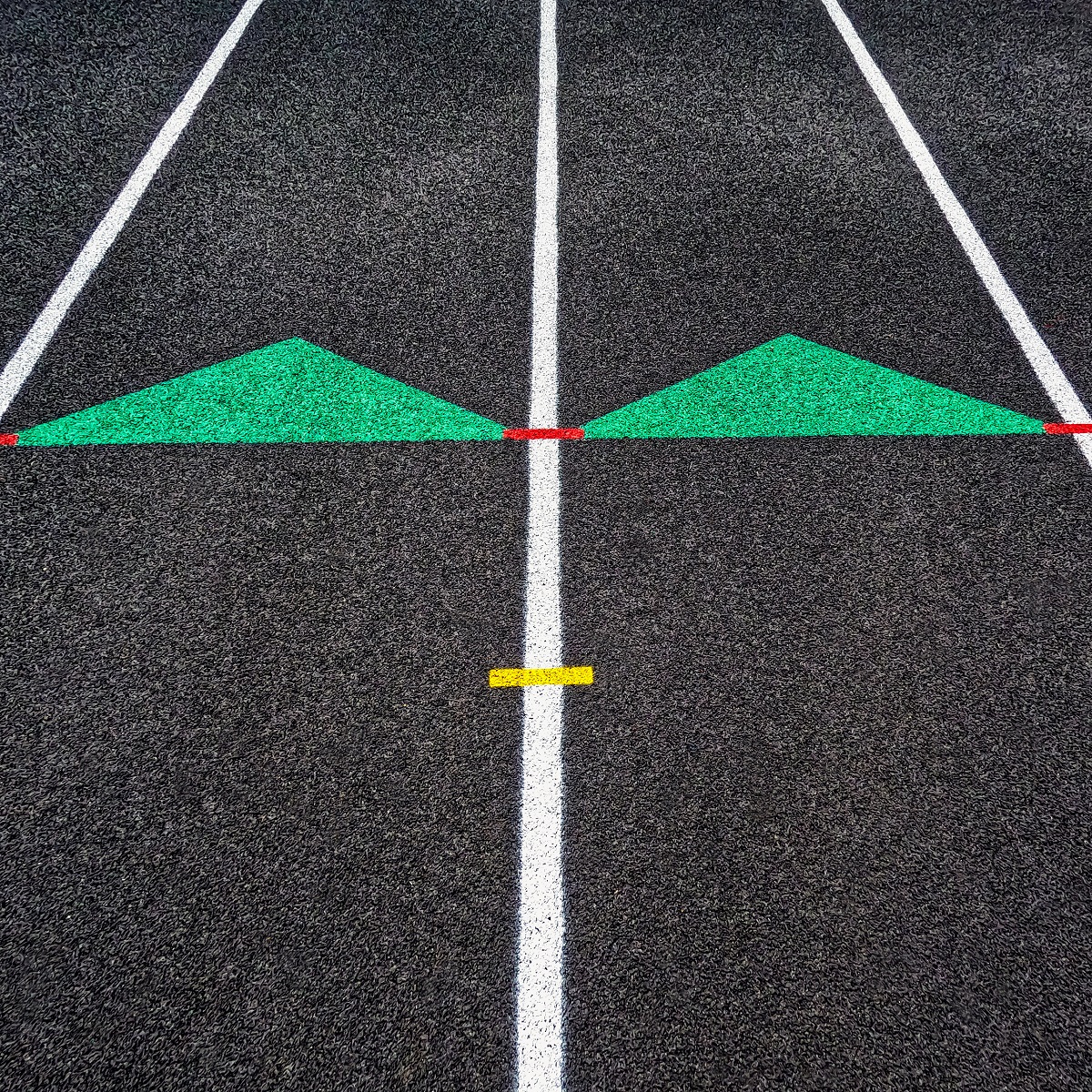 Overhead view of a rubber black running track surface with white lane lines. Green arrows point forward or upwards. Red dash marks connect the arrows. Yellow dash mark in the forefront of view. Richtungsmarkierung